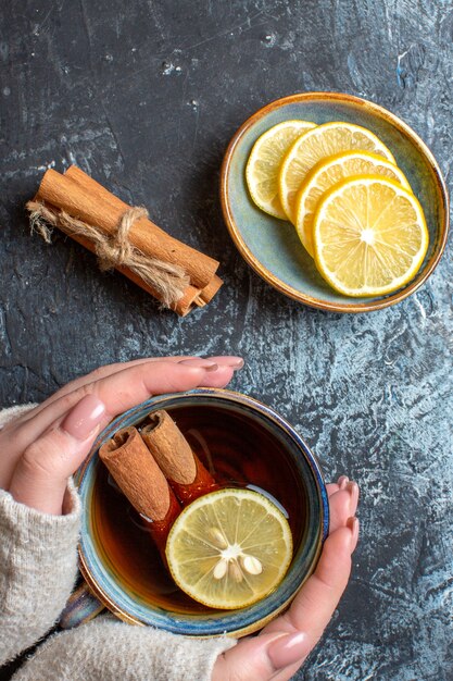 Vertikale Ansicht von frischen Zitronen und Hand, die eine Tasse schwarzen Tee mit Zimt auf dunklem Hintergrund hält