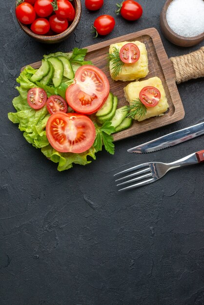 Vertikale Ansicht von geschnittenem frischem Tomaten- und Gurkenkäse auf Holzbrettbesteck stellte Salz auf schwarzer Oberfläche ein