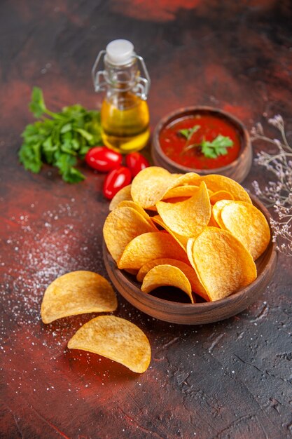 Vertikale Ansicht von hausgemachten köstlichen knusprigen Kartoffelchips in einer kleinen braunen Schüssel Ölflasche grüne Tomaten Knoblauchketchup auf dunklem Tisch