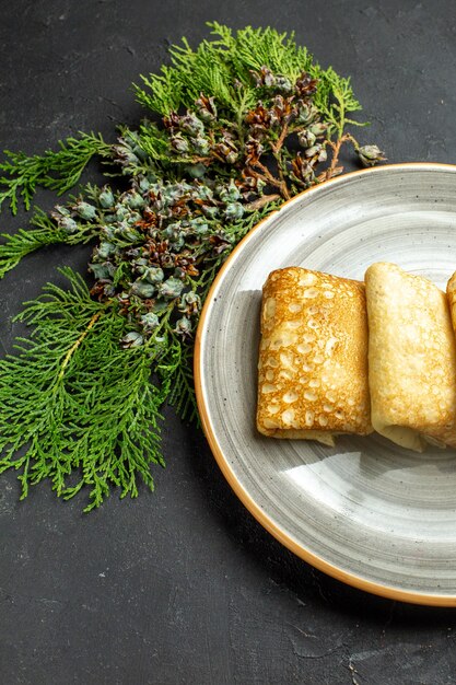 Vertikale Ansicht von köstlichen fleischgefüllten Pfannkuchen auf einem weißen Teller und einem Nadelbaumkegel auf schwarzem Hintergrund, halbes Schussfoto