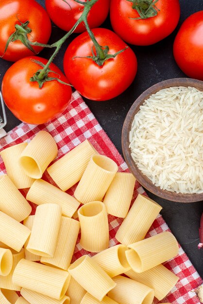 Vertikale Ansicht von rohen Nudeln auf rotem Handtuchreis in einer braunen Schüssel frische Tomaten auf schwarzem Tisch