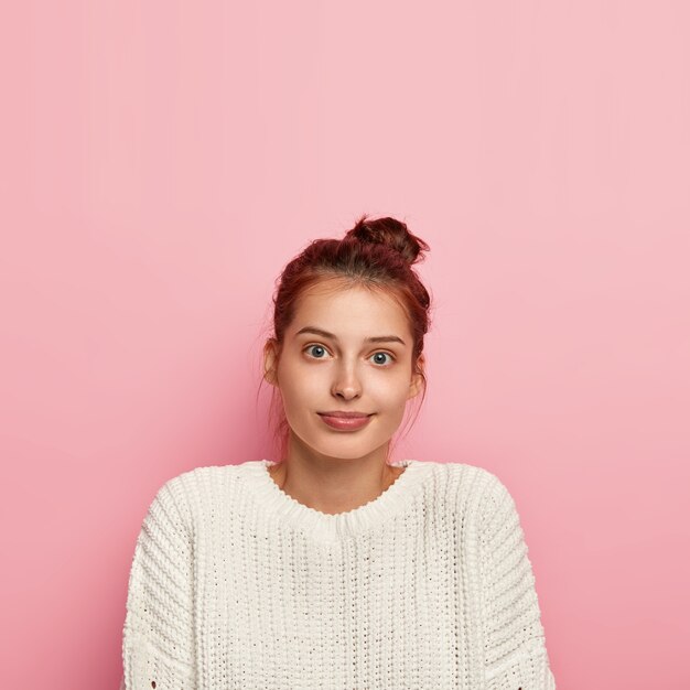 Vertikale Aufnahme des angenehm aussehenden zufriedenen tausendjährigen Mädchens trägt einen lässigen Strickpullover, hat volle Lippen und blaue Augen, dunkles Haar im Knoten gekämmt, posiert über rosa Hintergrund, Leerzeichen oben