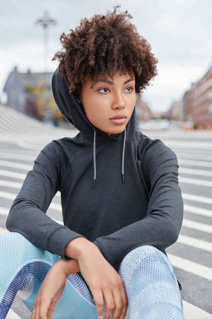 Kostenloses Foto vertikale aufnahme des attraktiven nachdenklichen afroamerikanischen hipsters trägt kapuzenpulli, sitzt auf asphalt