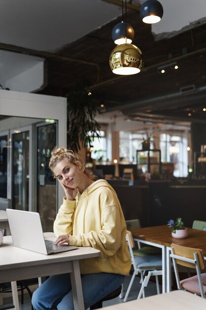 Vertikale Aufnahme des lächelnden attraktiven Mädchens, das in einem Café mit Laptop sitzt und Kamera betrachtet.