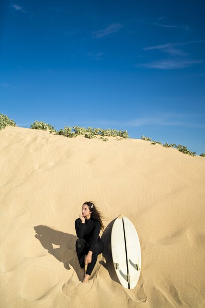 Vertikale Aufnahme einer attraktiven Frau, die auf einem sandigen Hügel mit einem Surfbrett auf der Seite sitzt