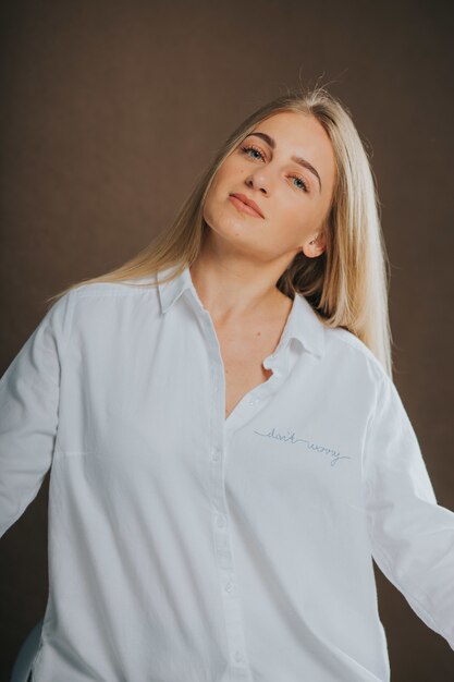 Vertikale Aufnahme einer attraktiven kaukasischen blonden Frau in einem weißen Hemd, die auf einer braunen Oberfläche posiert