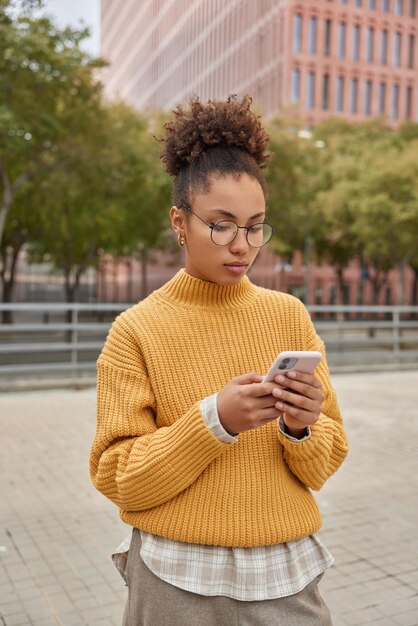 Vertikale Aufnahme einer aufmerksamen, lockigen jungen Frau trägt einen gelben Strickpullover, der auf dem Smartphone konzentriert ist