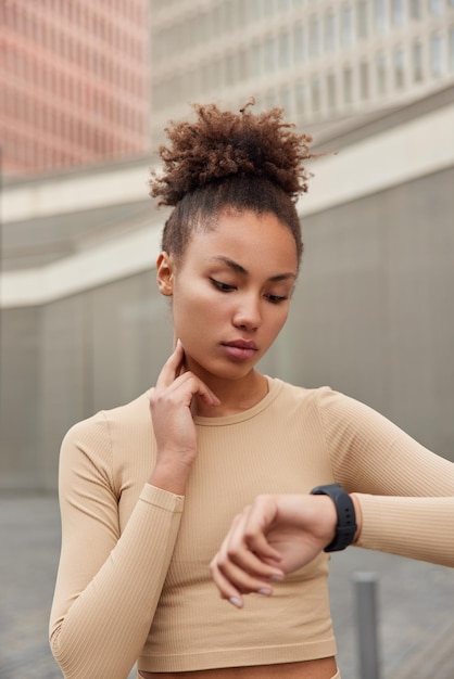 Vertikale Aufnahme einer ernsthaften Frau mit lockigen Haaren überprüft den Puls am Hals und konzentriert sich auf die in Sportbekleidung gekleidete Smartwatch, die die Herzfrequenz von Handgelenkshaltungen im Freien überwacht Sportliche Frau verwendet Trainingstracker