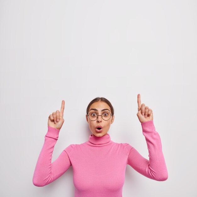 Vertikale Aufnahme einer erstaunten jungen Frau, die vor Verwunderung keucht, zeigt oben an, dass beide Zeigefinger eine optische Brille und einen lässigen rosa Polokragen tragen, der über weißer Wand isoliert ist?