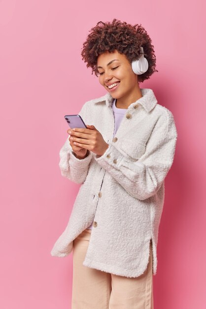 Vertikale Aufnahme einer fröhlichen Frau mit lockigem Haar hält Handy-Chats online und hört Lieblingsmusik in Kopfhörern trägt stilvolle Freizeitkleidung isoliert über rosa Wand