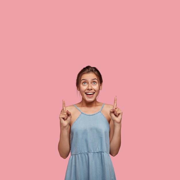 Vertikale Aufnahme einer fröhlichen jungen Frau mit fröhlichem Blick, zeigt nach oben, zeigt freien Raum, gekleidet in modisches Sommeroutfit
