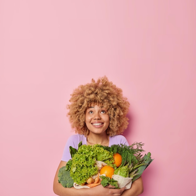 Kostenloses Foto vertikale aufnahme einer glücklichen, lockigen europäerin umarmt einen großen haufen grünes frisches gemüse, lächelt fröhlich und blickt isoliert über rosa hintergrund mit leerzeichen für ihre werbung