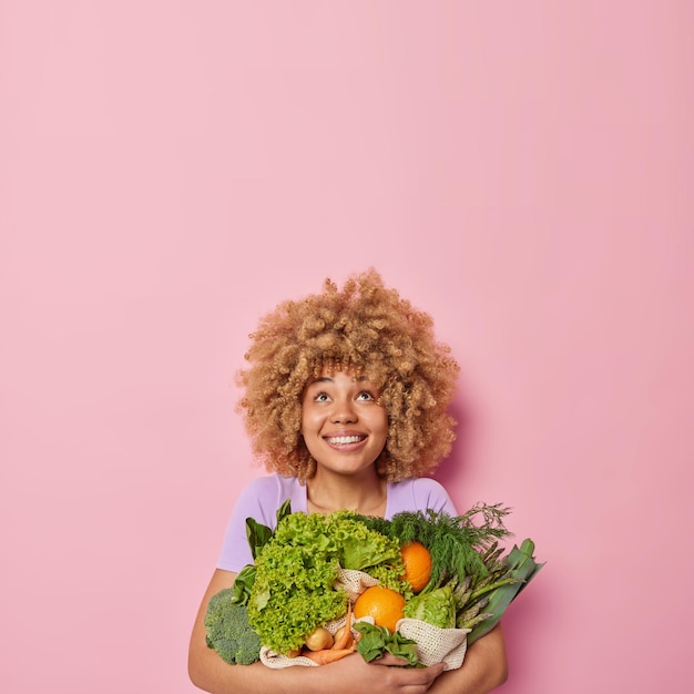 Vertikale Aufnahme einer glücklichen, lockigen Europäerin umarmt einen großen Haufen grünes frisches Gemüse, lächelt fröhlich und blickt isoliert über rosa Hintergrund mit Leerzeichen für Ihre Werbung