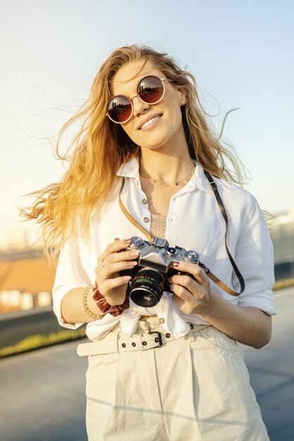 Vertikale Aufnahme einer glücklichen, modisch gekleideten Reisenden mit Kamera bei sonnigem Wetter