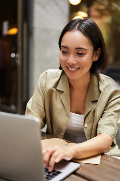 Vertikale Aufnahme einer jungen asiatischen Frau, die an der Fernbedienung von einem Café im Freien arbeitet, das mit Laptop und Smili sitzt