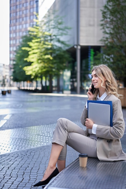 Vertikale Aufnahme einer jungen Unternehmensfrau mit Laptop und Arbeitsmappen, die am Handy sitzend spricht