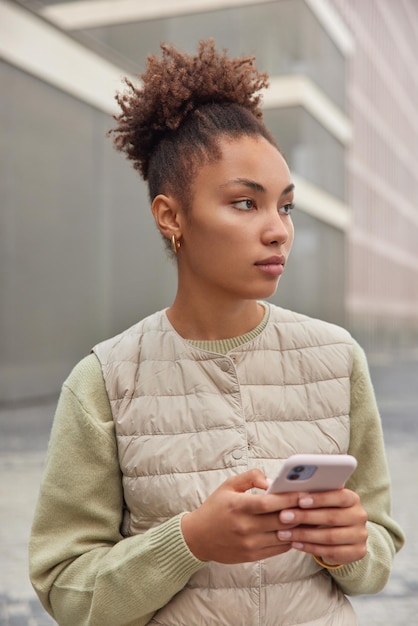 Kostenloses Foto vertikale aufnahme einer nachdenklichen frau mit lockigem, gekämmtem haar, gekleidet in einem lässigen pullover und einer weste, die das mobiltelefon verwendet, um neue anwendungen oder website-posen vor unscharfem hintergrund zu durchsuchen. moderne technologien