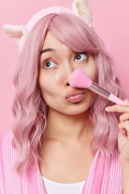 Vertikale Aufnahme einer nachdenklichen jungen asiatischen Frau, die einen Pinsel verwendet, trägt Make-up-Puder auf der Nase auf, hält die Lippen konzentriert weg, hat lange gefärbte Haare und trägt Stirnband-Posen im Innenbereich. Kosmetische Verfahren