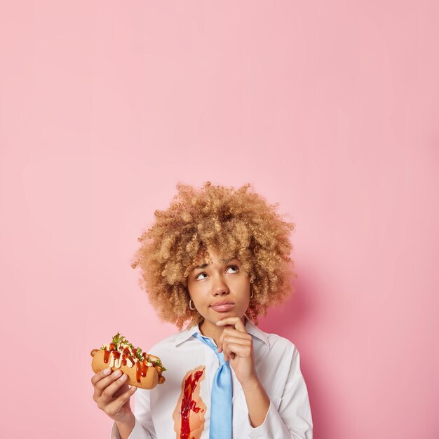 Vertikale aufnahme einer nachdenklichen lockigen frau isst leckeren hotdog hält das kinn konzentriert über dem kopf hat ungesunde ernährung trägt ein weißes hemd mit ketchup und eine blaue krawatte isoliert über einer rosa wand