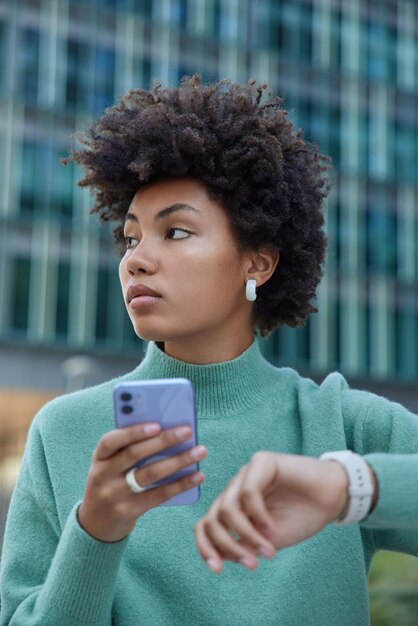 Vertikale Aufnahme einer nachdenklichen, lockigen jungen Frau, die darauf wartet, dass jemand die Zeit auf der Uhr überprüft, hält das Handy sendet SMS an einen Freund trägt lässige Pullover-Posen draußen vor unscharfem Hintergrund