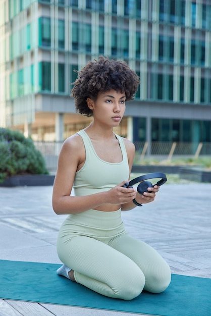 Vertikale aufnahme einer nachdenklichen sportlichen frau beim fitnesstraining im freien hält stereokopfhörer in activewear-posen auf knien bei karemat in der modernen stadt tagsüber gesundes lifestyle-konzept