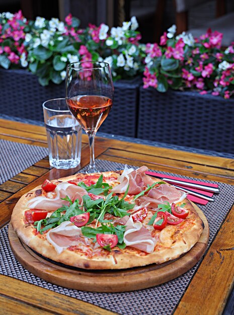 Vertikale Aufnahme einer Pizza mit Schinken und Tomaten auf einem Holzbrett auf dem Tisch mit Getränken darauf