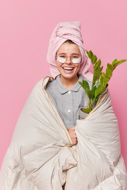 Vertikale Aufnahme einer positiven jungen Frau im Pyjama trägt Schönheitsflecken unter den Augen zur Hautbehandlung auf, die mit einer Decke umwickelt sind, nachdem sie geschlafen hat, hält eine Hauspflanze isoliert über rosa Hintergrund