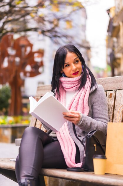 Vertikale Aufnahme einer schönen kaukasischen Frau, die auf einer Bank sitzt und ein Buch im öffentlichen Park liest