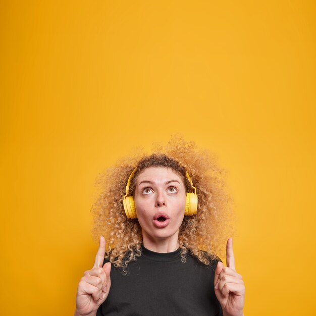 Vertikale Aufnahme einer überraschten, verwunderten jungen Frau mit lockigen Haarspitzen oben zeigt erstaunliche Werbung, die Musik über Kopfhörer hört, trägt ein lässiges gelbes T-Shirt isoliert über gelber Wand?