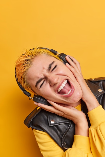 Vertikale Aufnahme einer weiblichen Hipster-Melomanin mit gefärbtem Haar kippt den Kopf, singt ein Lied, hört Musik in drahtlosen Kopfhörern trägt ein modisches Outfit und genießt eine Rock-Playlist, die über gelber Wand isoliert ist