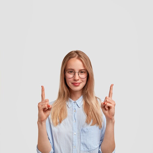 Vertikale Aufnahme einer zufriedenen jungen europäischen Frau mit erfreutem Ausdruck, zeigt mit Zeigefingern nach oben, gekleidet in modisches Hemd, zeigt etwas oben, isoliert über weißer Wand.