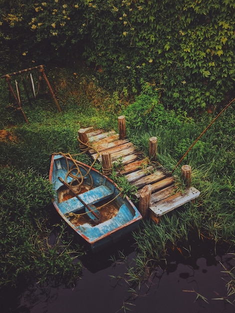 Kostenloses Foto vertikale aufnahme eines alten bootes im wasser nahe einem hölzernen dock, das durch grün umgeben ist