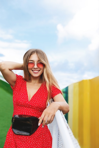 Vertikale Aufnahme eines hübschen blonden Mädchens im Sommerkleid und in der roten Sonnenbrille, die sich auf den Handlauf im Park stützen.