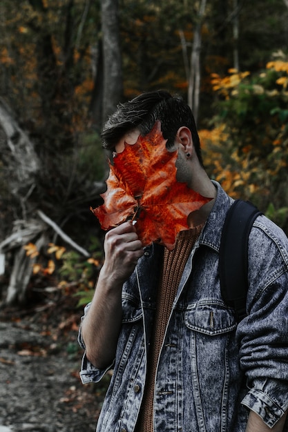 Vertikale Aufnahme eines jungen Mannes, der sein gesamtes Gesicht mit einem großen goldenen Herbstblatt bedeckt