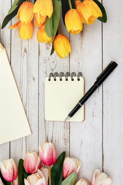 Vertikale Aufnahme eines leeren Notizbuchs und eines Stifts, einige Blumen auf einer Holzoberfläche