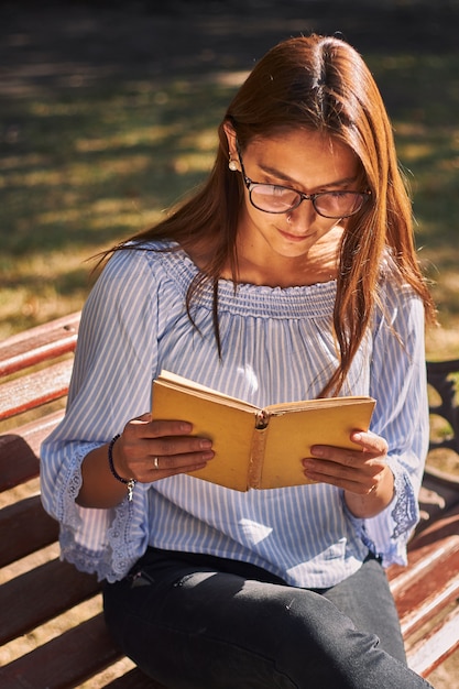 Vertikale Aufnahme eines Mädchens in einem blauen Hemd und in der Brille beim Lesen eines Buches auf der Bank