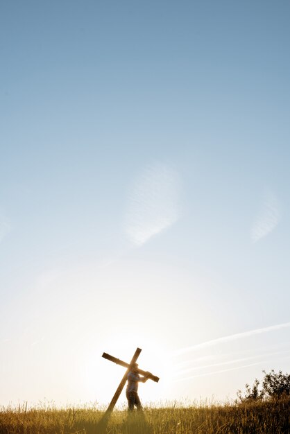 Vertikale Aufnahme eines Mannes, der ein großes Holzkreuz in einem Grasfeld mit blauem Himmel trägt