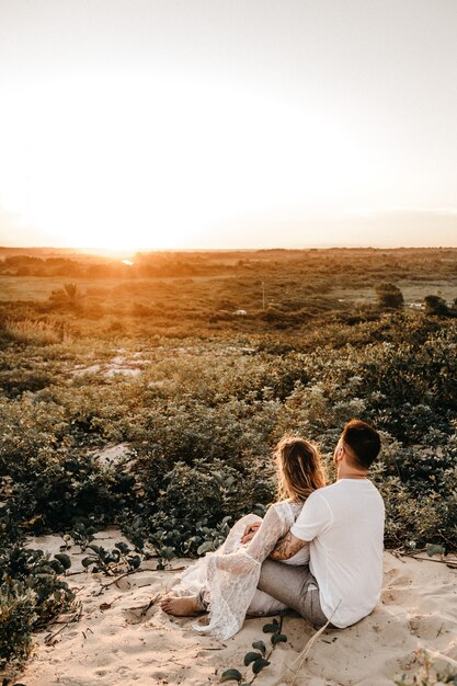 Vertikale Aufnahme eines Mannes und einer Frau, die in einem Feld sitzen und sich umarmen, während sie den Sonnenuntergang betrachten
