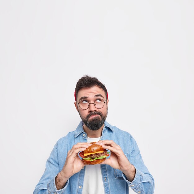 Vertikale Aufnahme eines nachdenklichen bärtigen Mannes, der einen appetitlichen Hamburger hält, der sich oben konzentriert, denkt tief über etwas nach