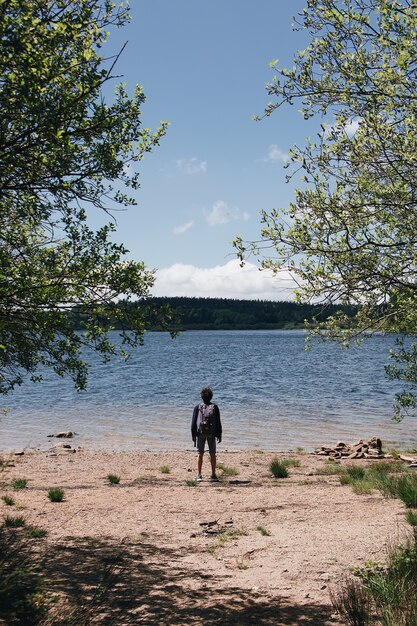 Vertikale Aufnahme eines Wanderers, der am Strand an einem See und den Hügeln im Hintergrund steht