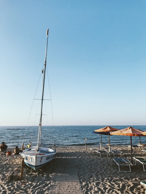 Kostenloses Foto vertikale aufnahme eines weißen segelboots am ufer in der nähe des wassers mit einem blauen himmel in der