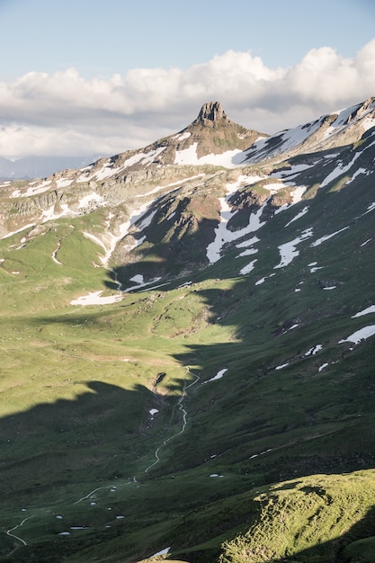 Vertikale Aufnahme von grasbewachsenen Hügeln nahe einem schneebedeckten Berg mit einem bewölkten Himmel