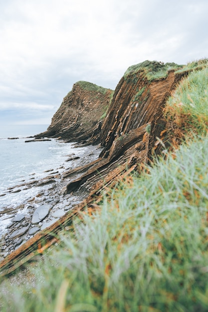 Vertikale Aufnahme von Klippen gefüllt mit grünem Gras neben dem blauen Meer bei Tageslicht