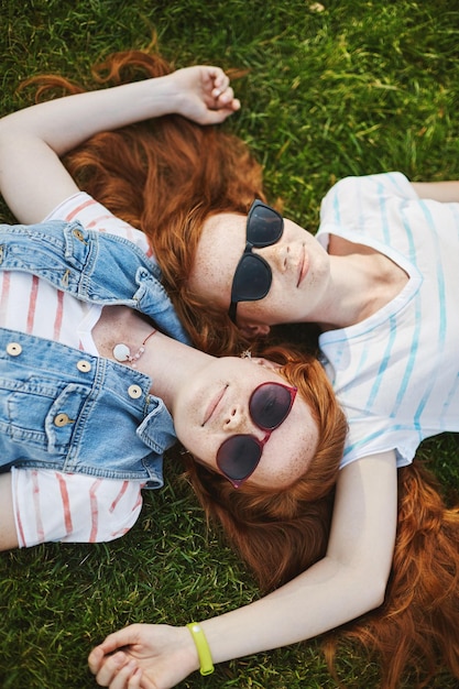 Vertikale Aufnahme von schönen, entspannten und glücklichen Mädchen mit natürlichem Ingwerhaar und süßen Sommersprossen, die auf Gras liegen und sich gegenseitig berühren, tragen coole Sonnenbrillen, die an warmen Sommertagen unter der Sonne chillen