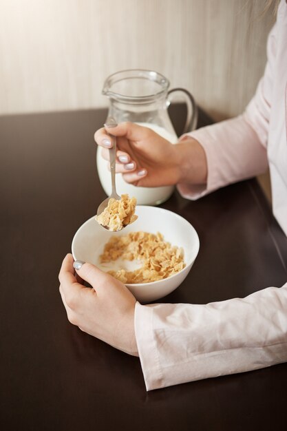 Vertikale beschnittene Aufnahme der Frau, die in der Küche sitzt und Löffel hält, während sie Schüssel Müsli mit Milch isst, gesundes Frühstück hat und schönen Morgen mit Familie genießt, Pläne für heute besprechend