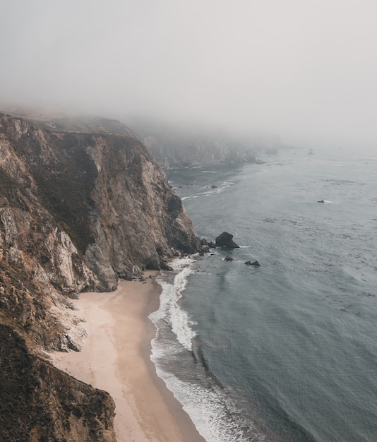 Vertikale Luftaufnahme einer Klippe am Meer mit sandigem Ufer unter einem nebligen Himmel