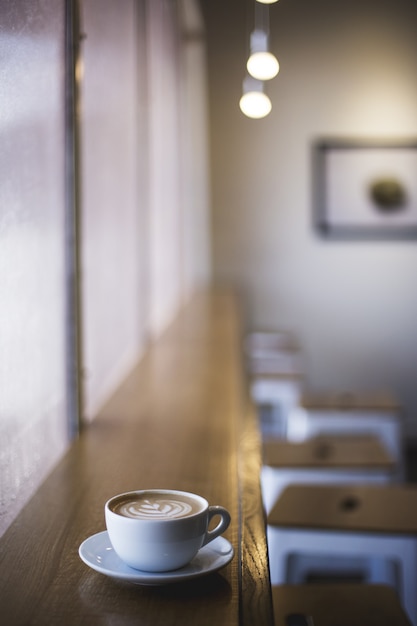 Vertikale Nahaufnahmeaufnahme des weißen Bechers des Latte-Kunstkaffees auf einem Fensterregal in einem Café