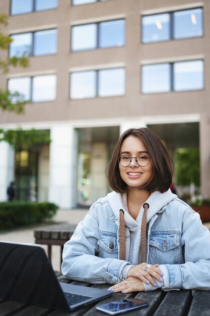 Vertikale Porträt intelligente gut aussehende Studentin in Brille Jeansjacke, sitzen draußen auf der Bank, arbeiten im Park, Teilzeit freiberuflicher Job während des Studiums in der Universität, Laptop und Handytisch.