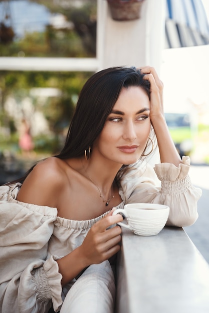 Vertikaler Schuss der schönen brünetten Frau mit goldener Bräune, sitzend an einem Café mit einer Tasse Kaffee und Blick auf Straße.