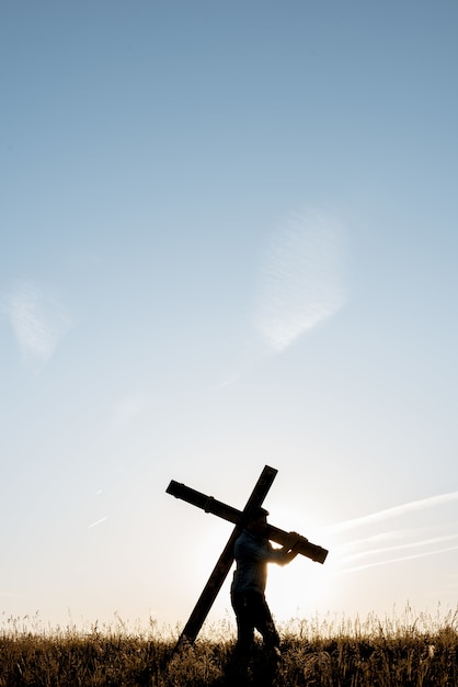 Vertikaler Schuss eines Mannes, der ein handgemachtes Holzkreuz in einem Grasfeld unter einem blauen Himmel trägt